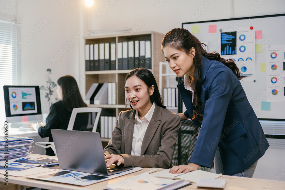 Poster two asian businesswomen are working together, using a laptop in a modern office to discuss a project