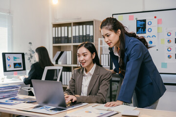 Two asian businesswomen are working together, using a laptop in a modern office to discuss a project, with charts and graphs visible on the screen and printed documents on the desk