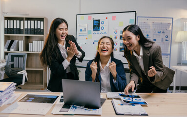 Three young asian businesswomen are celebrating a successful project, laughing and cheering with fists raised in front of a laptop and documents in a modern office