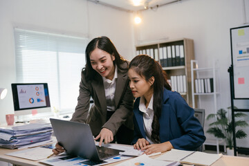 Two smiling asian businesswomen are collaborating on a laptop, analyzing charts and financial data, demonstrating teamwork and expertise in a modern office environment