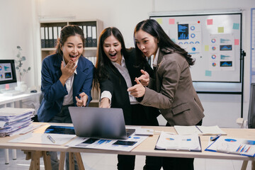 Three excited asian businesswomen are pointing at a laptop screen, celebrating a successful business achievement in their modern office