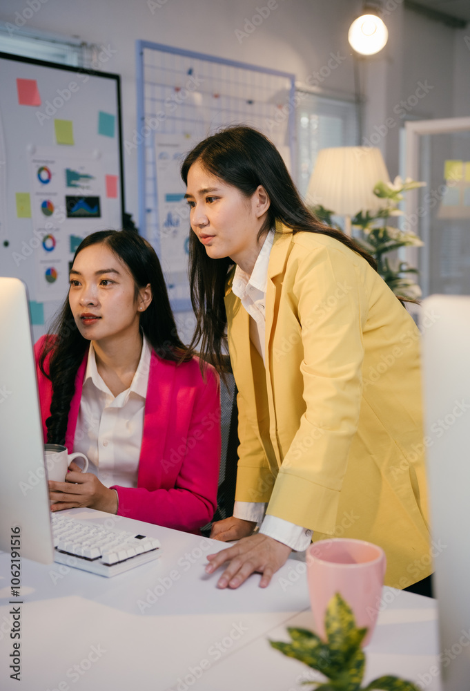 Poster Two young businesswomen wearing colorful blazers are collaborating on a project, focusing on a desktop computer in a contemporary office setting with charts and notes visible in the background