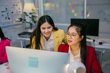 Two asian businesswomen are working together in a modern office, collaborating on a computer project, with one offering guidance and support to the other