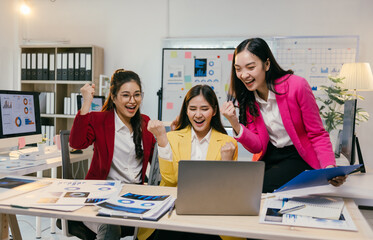 Three young businesswomen are cheering and raising their fists in celebration of a successful business achievement while working together on a laptop in a modern office