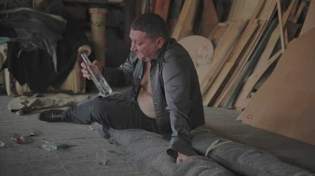 A very drunk man lies on cement with a bottle of vodka.A drunk man drinks vodka in his workshop.Concept of alcoholism. Man drinking vodka alone in a dark room in slow-motion.