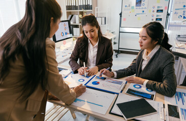Three asian businesswomen are discussing and analyzing financial charts and graphs during a productive meeting in a modern office, demonstrating teamwork and collaboration