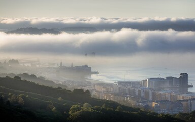 Naklejka premium A city shrouded in fog at sunrise, with distant buildings and a coastline visible through the mist.