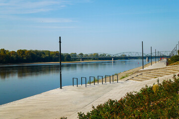 Waterfront with blue skies, a concrete walkway with metal railings and lights leading to a metal bridge in the distance.