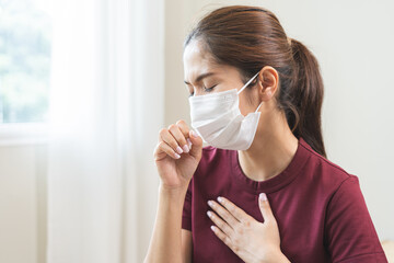 Unhealthy sickness asian young woman wearing face mask, unwell coughing, have cold, sore throat on sofa at home, suffering with symptom cough feeling bad. Health care of Coronavirus, covid-19 concept.