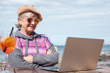 Smiling senior woman using laptop computer in outdoor at the beach in a sunny day enjoying a fresh drink. Portrait of elderly lady in vacation or retirement at sea