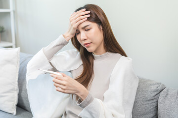 Sick, influenza asian young woman, girl headache have fever, hand touching forehead, holding thermometer for check measure body temperature, illness while sitting rest on sofa at home. Health care.