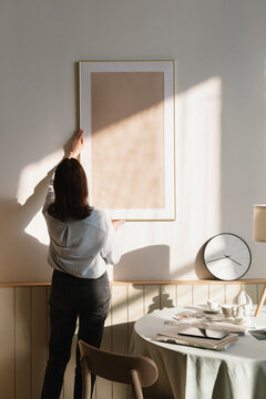 Rear view of a woman hanging a blank picture frame on a  wall in a living room