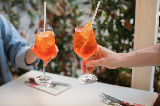 Close-up of a man and woman at an outdoor restaurant clinking glasses with orange spritz cocktails