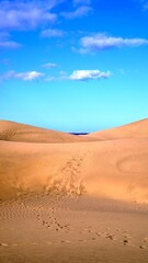 Texture and footprints on desert sand with dunes in the background. Blue sky with clouds, islands and ocean on the horizon