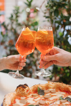 Close-up of a man and a woman clinking glasses with orange spritz cocktails and eating traditional Italian sausage pizza