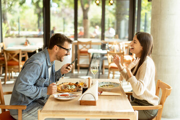 Couple enjoying a cozy meal together in a bright cafe, capturing memories with a smartphone during a sunny afternoon