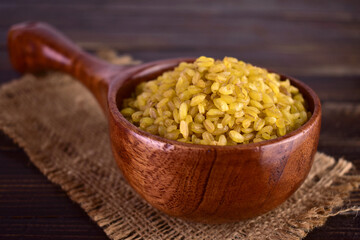 Bulgur cereal in a wooden spoon on a dark background.
