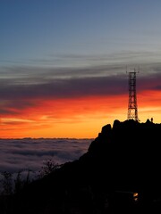 A tall cell tower stands silhouetted against a vivid sunset, its outline dramatically set against a backdrop of rolling clouds and brightly colored sky - Pico do Arieiro, Madeira, Portugal.