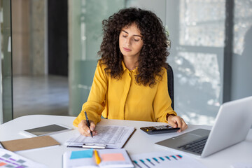 Businesswoman in yellow blouse focused on calculations using documents and calculator at office desk with laptop and charts. Represents productivity, finance, and professional work setting.