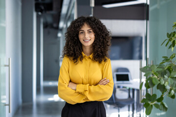 Confident businesswoman with curly hair in yellow blouse stands with arms crossed in modern office...