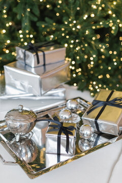 Close-up of a stack of wrapped gifts in silver paper on a silver tray next to an illuminated Christmas tree