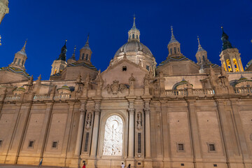 Fototapeta premium Illuminated Basilica del Pilar in Zaragoza, Spain, at dusk, with serene reflections on the Ebro River. Ideal for travel, architecture, and cultural themes.