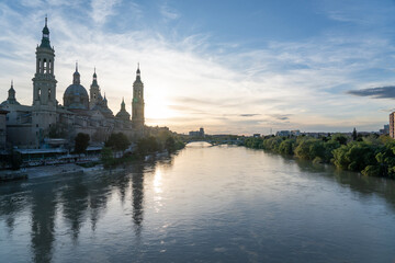 Obraz premium Illuminated Basilica del Pilar in Zaragoza, Spain, at dusk, with serene reflections on the Ebro River. Ideal for travel, architecture, and cultural themes.