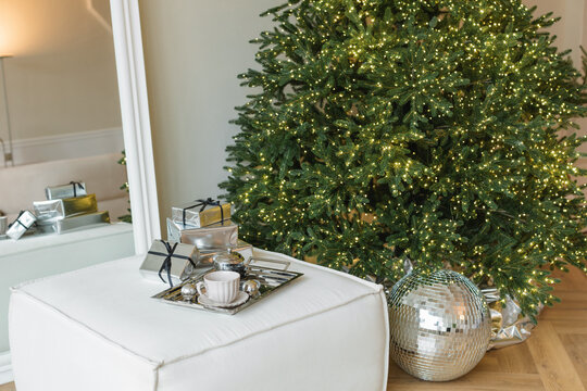 Stack of wrapped Christmas gifts on a leather cube pouffe next to an illuminated Christmas tree and glitter ball in a stylish living room