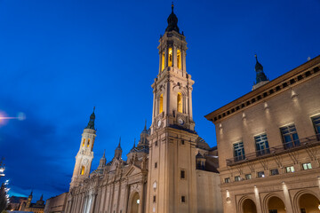 Fototapeta premium Illuminated Basilica del Pilar in Zaragoza, Spain, at dusk, with serene reflections on the Ebro River. Ideal for travel, architecture, and cultural themes.