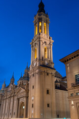 Fototapeta premium Illuminated Basilica del Pilar in Zaragoza, Spain, at dusk, with serene reflections on the Ebro River. Ideal for travel, architecture, and cultural themes.
