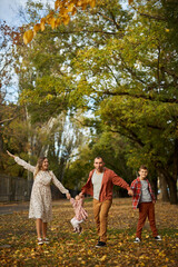 Happy family walking on green grass strewn with fallen leaves in Park in autumn. Parents, two children are walking in park at sunset with holding hands. Family, child, walk through Autumn Park.