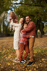 Happy family walking on green grass strewn with fallen leaves in Park in autumn. Parents, two children are walking in park hug and laugh. Family, child, walk through Autumn Park.