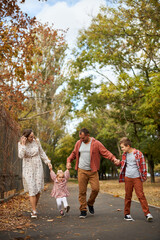 Fototapeta premium Happy family walking on green grass strewn with fallen leaves in Park in autumn. Parents, two children are walking in park at sunset with holding hands. Family, child, walk through Autumn Park.