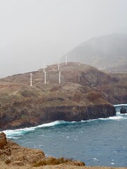 The image showcases wind turbines on a foggy hill, surrounded by sparse greenery and a desert-like landscape, representing renewable energy in a barren environment - Madeira, Portugal.