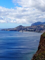 A breathtaking view of a coastal city nestled between the ocean and mountains, with clouds overhead, symbolizing harmony, grandeur, and nature magnificence - Madeira, Portugal.