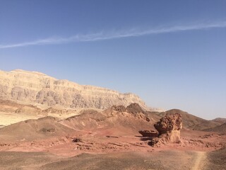 The photo depicts a vast, rugged desert landscape in Timna Valley with brown terrain and rock formations under a clear blue sky. In the background, steep cliffs add depth to this serene, arid view.