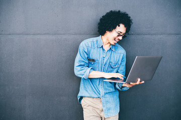 Young man with curly hair wearing a blue denim shirt stands against a dark gray wall, holding a laptop in one hand and typing with the other, smiling and looking focused on his work