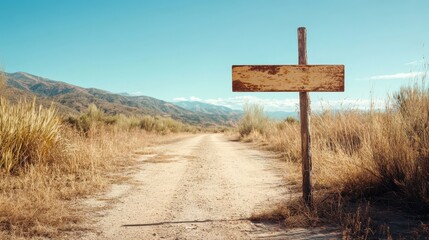 Naklejka premium An aged, rectangular wooden sign post stands by an unpaved dirt road amidst dry grasses, leading towards a distant mountainous horizon under a clear blue sky.