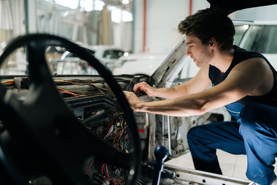 Focused mechanic in overalls examining the car electrical system in repair shop, with focus on dashboard wiring. Concept of professional car repair and maintenance work.