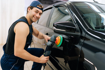 Portrait of positive mechanic male in uniform polish auto door using grinding unit in automobile repair and renew service station, smiling looking at camera. Concept of car repair and maintenance work