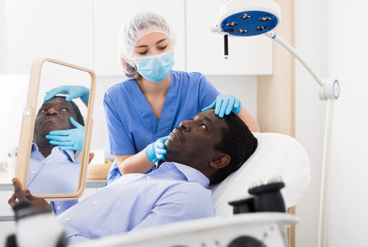 Skilled female beautician in face mask giving consultation to african male patient in aesthetic clinic
