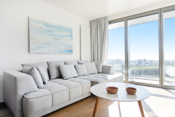 Cozy interior of modern living room with large panoramic window, comfy grey sofa, stylish coffee table, room in white and grey soft colors