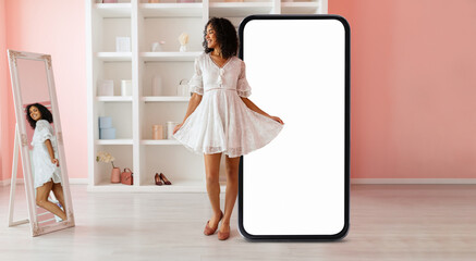 A young black woman wearing a stylish white dress stands joyfully next to an oversized blank phone display, with a mirror reflecting her image in a chic, softly colored room.