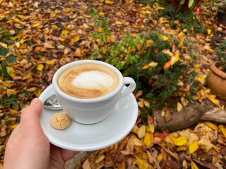 White cup with hot coffee drink in white female hand on blurry background of autumn garden with yellow fallen leaves outside