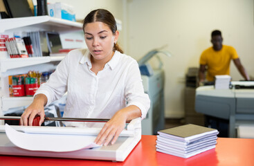 Concentrated young woman in white shirt using paper cutter on the table with planners and...