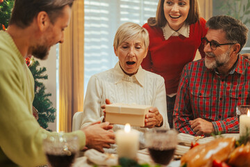Family having christmas dinner giving gift to surprised senior woman