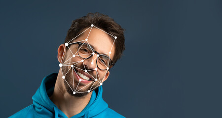 A young man with glasses smiles brightly while wearing a hoodie. The background is dark, highlighting facial recognition markers on his face, illustrating advances in technology.