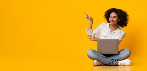 Nice Promo. Happy Black Woman With Laptop On Lap Pointing At Copy Space Over Yellow Background, Sitting On Floor In Studio, Panorama