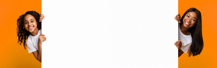 Promotion Billboard Concept. Cheerful African Woman Mom And Girl Holding White Advertisement Board, Standing Together On Yellow Background, Looking At Camera Peeping Out The Two Sides Of Sign Board