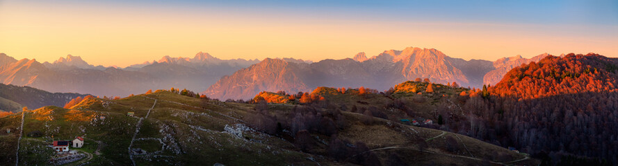 Naklejka premium The golden hues of sunset illuminate the mountainous landscape from Pizzoc mountain toward Dolomites, highlighting the rolling hills and serene valleys below, showcasing nature's beauty at dusk.